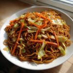 A close-up of a white bowl filled with savory fried noodles mixed with bright orange julienned carrots and wilted cabbage.