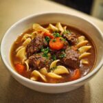 Close-up of a white bowl filled with rich beef noodle soup, featuring tender beef chunks, egg noodles, and sliced carrots.