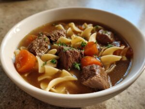Close-up of a white bowl filled with amazing beef noodle soup, featuring chunks of beef, egg noodles, and carrots.