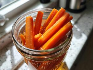 Close-up of bright orange pickled carrots sticks submerged in brine inside a clear glass jar.