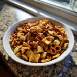 A white bowl filled with seasoned Chex party mix, featuring square cereal pieces and small pretzels, sitting on a granite counter.