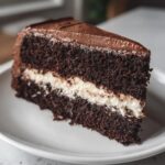A tempting slice of dark chocolate cake with a thick white cream filling and chocolate frosting, reminiscent of an italian love cake.