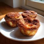 Three freshly baked cinnamon sugar french toast muffins coated in a thick cinnamon sugar crust, served on a white plate.