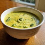A close-up of a white bowl filled with thick, creamy broccoli cheddar soup, showing florets and melted cheese swirls.