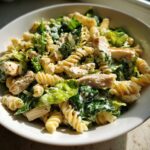 A close-up, bright photo of a bowl filled with creamy chicken caesar pasta salad featuring rotini pasta, grilled chicken pieces, and romaine lettuce.