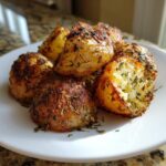 A close-up of golden, crispy oven roasted potatoes seasoned generously with dried herbs on a white plate.
