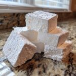 A stack of freshly cut, fluffy homemade marshmallows generously dusted with powdered sugar, resting on a granite countertop.