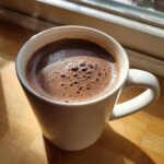 A close-up of a steaming mug of rich, dark hot cocoa mix with small bubbles on the surface, sitting on a wooden counter in sunlight.