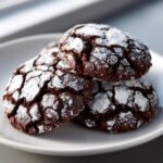 A stack of three rich, dark chocolate crinkle cookies dusted heavily with powdered sugar.