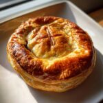 A golden-brown, perfectly baked brie in puff pastry resting on a white plate, glistening under sunlight.