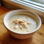A close-up of a white bowl filled with creamy homemade cream of chicken soup, topped with shredded chicken and herbs.