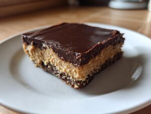 A close-up of a layered peanut butter bars slice with a chocolate topping on a white plate.