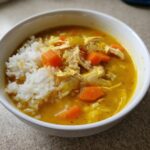 A close-up of a white bowl filled with vibrant yellow Lemon Ginger Turmeric Chicken and Rice Soup, showing shredded chicken, carrots, and white rice.