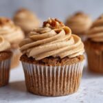 A close-up of one perfectly frosted pecan pie cupcake topped with chopped pecans, surrounded by more cupcakes.