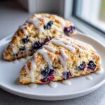 Two triangular blueberry scones drizzled generously with white vanilla glaze, sitting on a white plate.