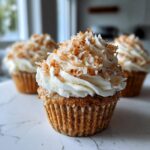 A close-up of a moist coconut cupcake topped with thick white frosting and toasted coconut flakes. More coconut cupcakes are blurred in the background.