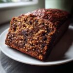 Close-up of a moist slice of dark, rich date nut bread served on a white plate.