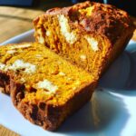 A loaf of moist pumpkin cream cheese bread, sliced to show the bright orange interior and swirls of white cream cheese.