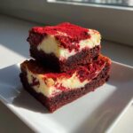 Two stacked, rich red velvet cheesecake brownies dusted with red powder on a white plate.