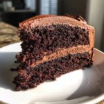 A close-up, mouthwatering slice of multi-layered chocolate cake with thick chocolate frosting, sitting on a white plate.