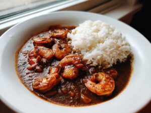 A close-up of a bowl of rich, dark shrimp etouffee served alongside a mound of white rice.