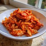 Close-up of flavorful shredded chicken tinga coated in a vibrant red, savory sauce, served in a white bowl.