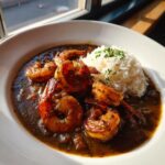 Close-up of a bowl of dark, rich shrimp etouffee featuring large seasoned shrimp next to a scoop of white rice.