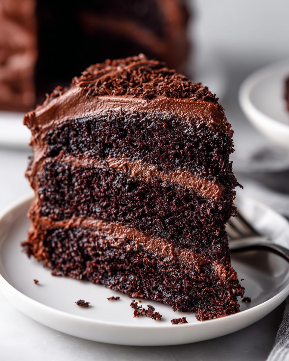 Close-up of a moist slice of three-layer chocolate cake with rich chocolate frosting, topped with chocolate shavings.