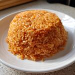 A perfectly molded mound of vibrant orange smoky jollof rice served on a white plate.