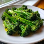 Close-up of bright green steamed broccoli florets and stalks tossed in olive oil and herbs on a white plate.