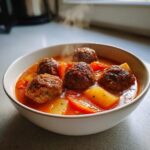 Close-up of a steaming white bowl filled with rich, red albondigas soup containing large meatballs, potatoes, and carrots.