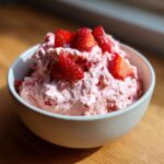 A close-up of a bowl filled with light pink, fluffy strawberry cheesecake dip, topped with fresh, sliced strawberries.