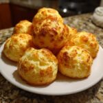 A mound of freshly baked, golden-brown Brazilian cheese bread rolls stacked on a white plate.