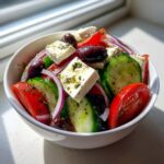 Close-up of a fresh greek salad in a white bowl featuring feta cheese cubes, cucumbers, tomatoes, olives, and red onion.