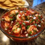 Close-up of a glass bowl filled with vibrant mexican shrimp cocktail, featuring plump shrimp, tomatoes, onions, and cilantro.