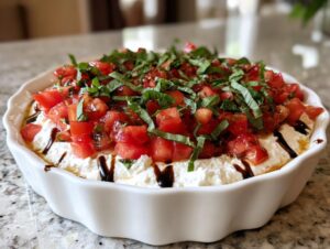 A white fluted dish filled with creamy bruschetta dip topped with diced tomatoes, fresh basil, and a balsamic glaze drizzle.