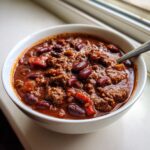Close-up of a white bowl filled with rich, thick chili featuring ground meat and kidney beans.