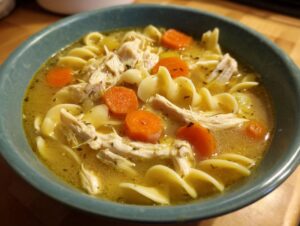 Close-up of a teal bowl filled with homemade chicken soup, featuring shredded chicken, egg noodles, and bright orange carrots.