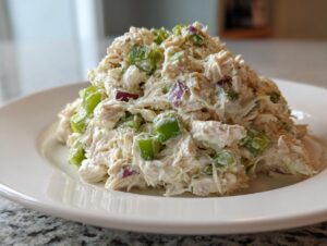 A mound of creamy chicken salad featuring shredded chicken, celery, and red onion on a white plate.