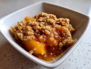 Close-up of a single serving of warm peach crisp with bubbly fruit filling and crunchy topping in a white bowl.