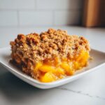 A close-up of a square serving of homemade peach crisp showing gooey peach filling and a thick, golden brown oat topping.