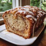 A loaf of freshly baked apple fritter bread drizzled with white vanilla glaze, served on a white plate.
