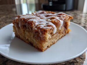 A thick, square slice of apple pie bars topped with a white vanilla glaze, served on a white plate.