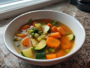 Close-up of a white bowl filled with bright vegetable soup featuring sliced carrots, zucchini, and peas.