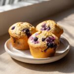 Three golden-brown blueberry cottage cheese muffins sitting on a small white plate near a window.