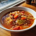 A close-up of a white bowl filled with rich, reddish broth, chunks of cabbage, carrots, and ground beef, representing delicious cabbage soup.