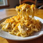 A serving of creamy, cheesy chicken spaghetti with visible chunks of chicken, being lifted by a fork showing long cheese pulls.