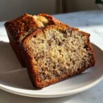 A thick slice of moist banana bread with visible chocolate chips sits next to the rest of the loaf.