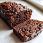 A close-up of a loaf of rich, dark chocolate zucchini bread with a slice cut, topped with melted chocolate chips.