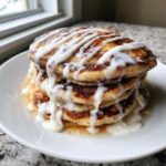 A tall stack of fluffy cinnamon roll pancakes drizzled generously with white cream cheese icing.